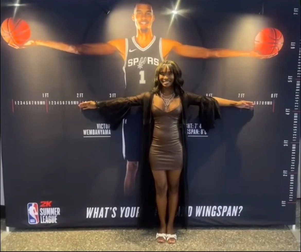 Woman standing in front of a basketball-themed measuring wall, with her arms stretched out, mimicking a basketball player with a big smile, in front of a large image of a basketball player in a San Antonio Spurs jersey.