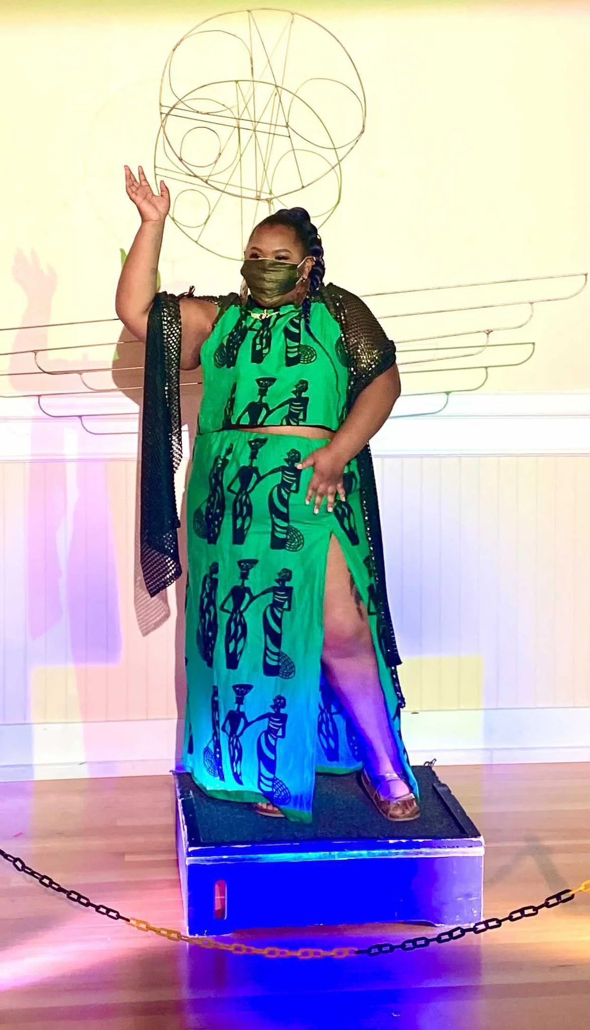 Black, female model wearing a green dress with black figures patterened  on it. She stands on a small pedestal with one arm stretched above her showcasing a delicate, circular metal sculpture above her head.