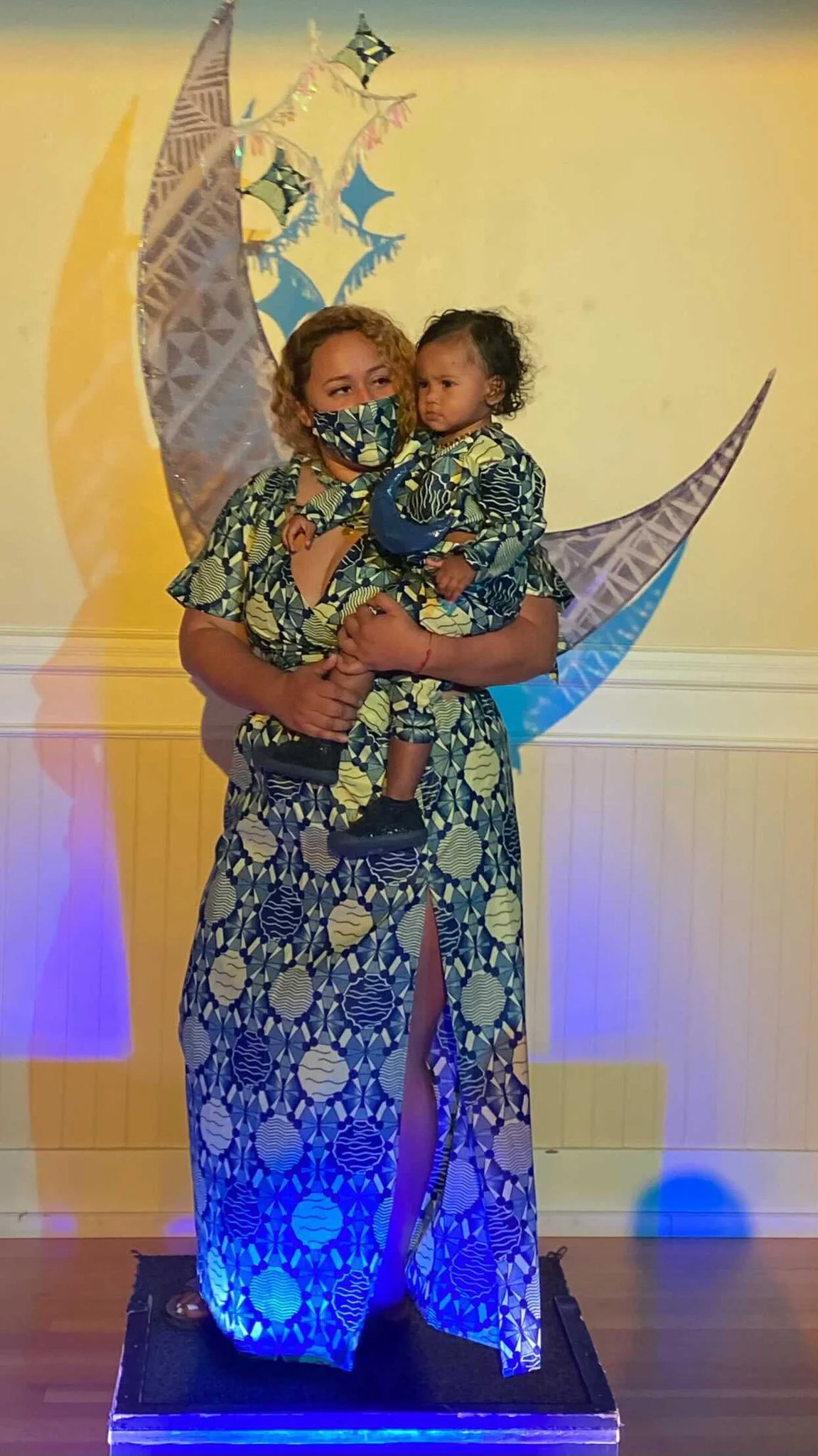 A black, female model holds a child in her arms. They both wear matching blue and silver patterned clothing and a crescent moon shaped sculpture stretches behind them