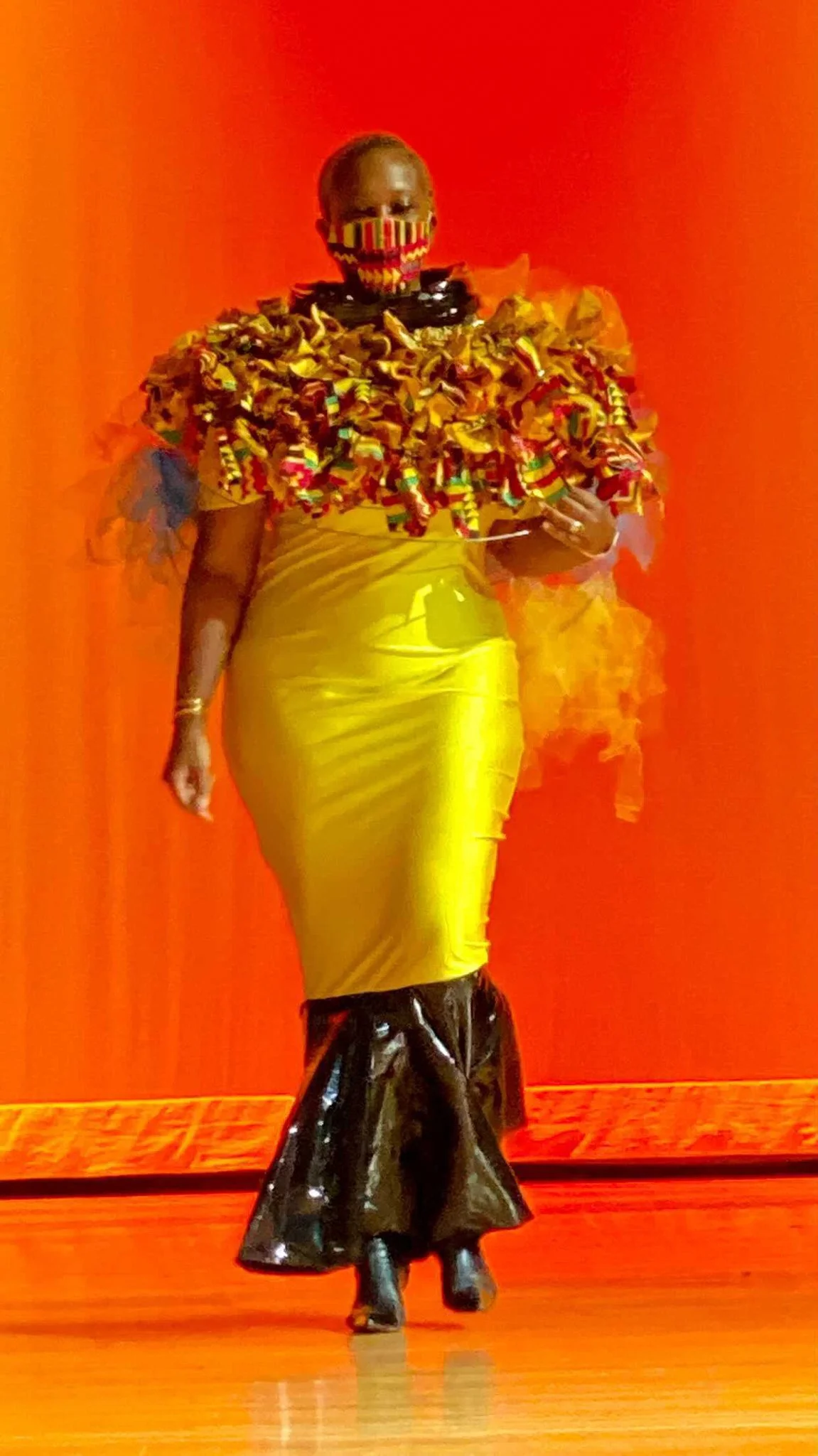 A female, black model in a yellow mermaid- dress with elaborate ruched color and black fins at the bottom. 