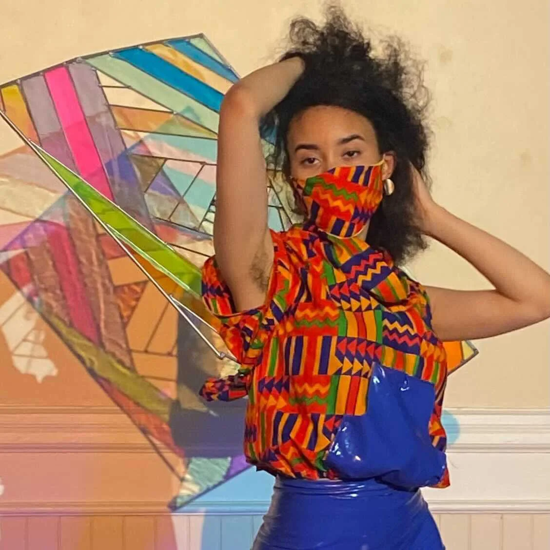 Close up of a black, female model wearing an orange patterned sleeveless top with matching mask. A delicate rectangular sculpture extends from her back.