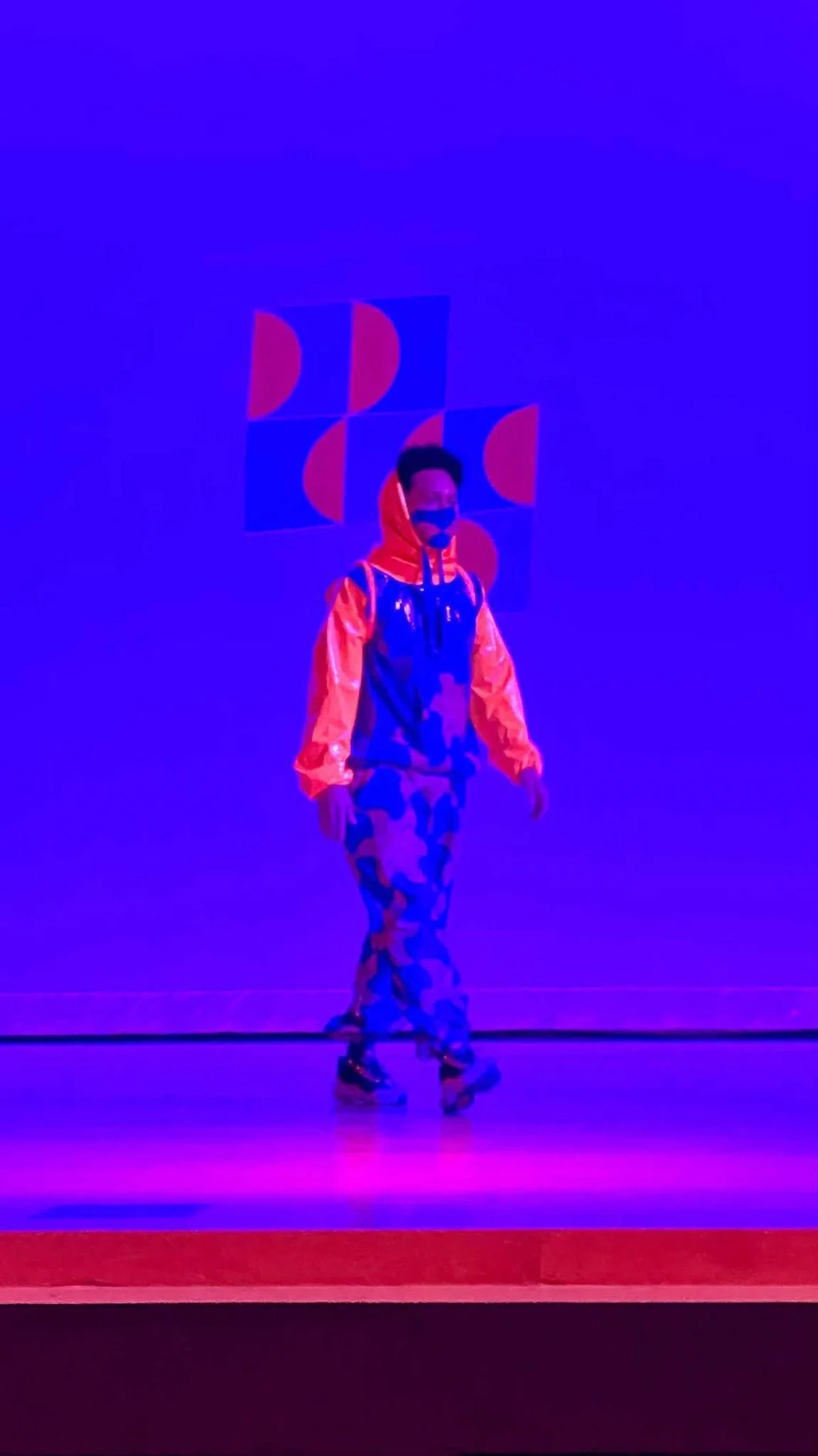 a model on the runway in a long blue and orange garment with an orange hoodie and wide sleeves