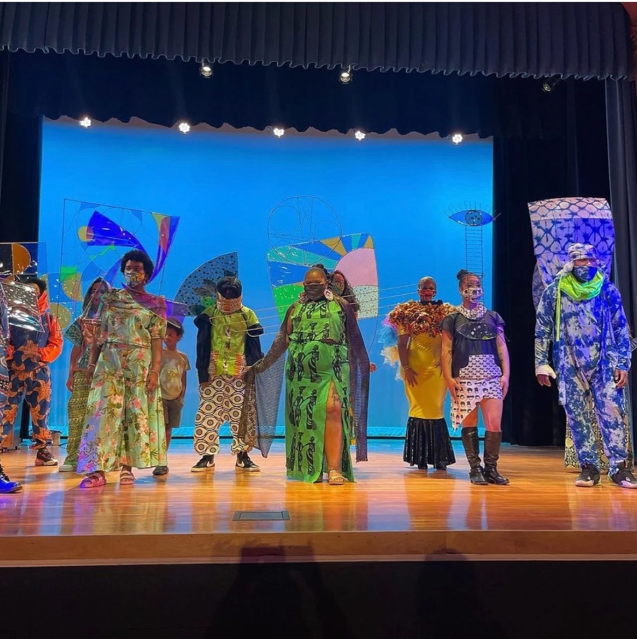 A group of nine models standing on a stage all wearing colorful and elaborate garments