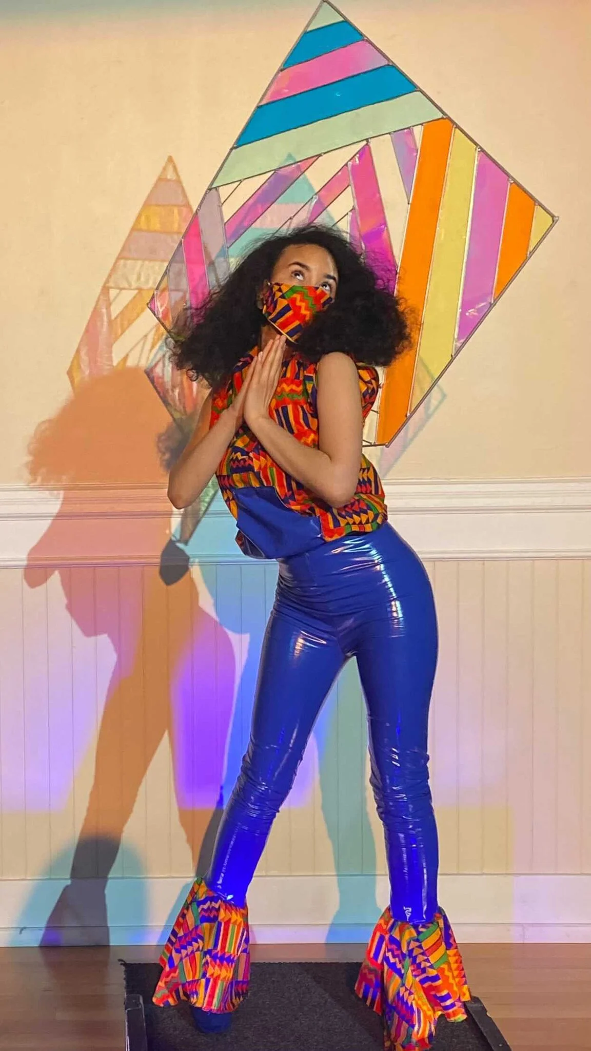 A black, female model wears an orange patterned sleeveless top with matching mask over form fitting blue pants. A delicate rectangular sculpture extends from her back.