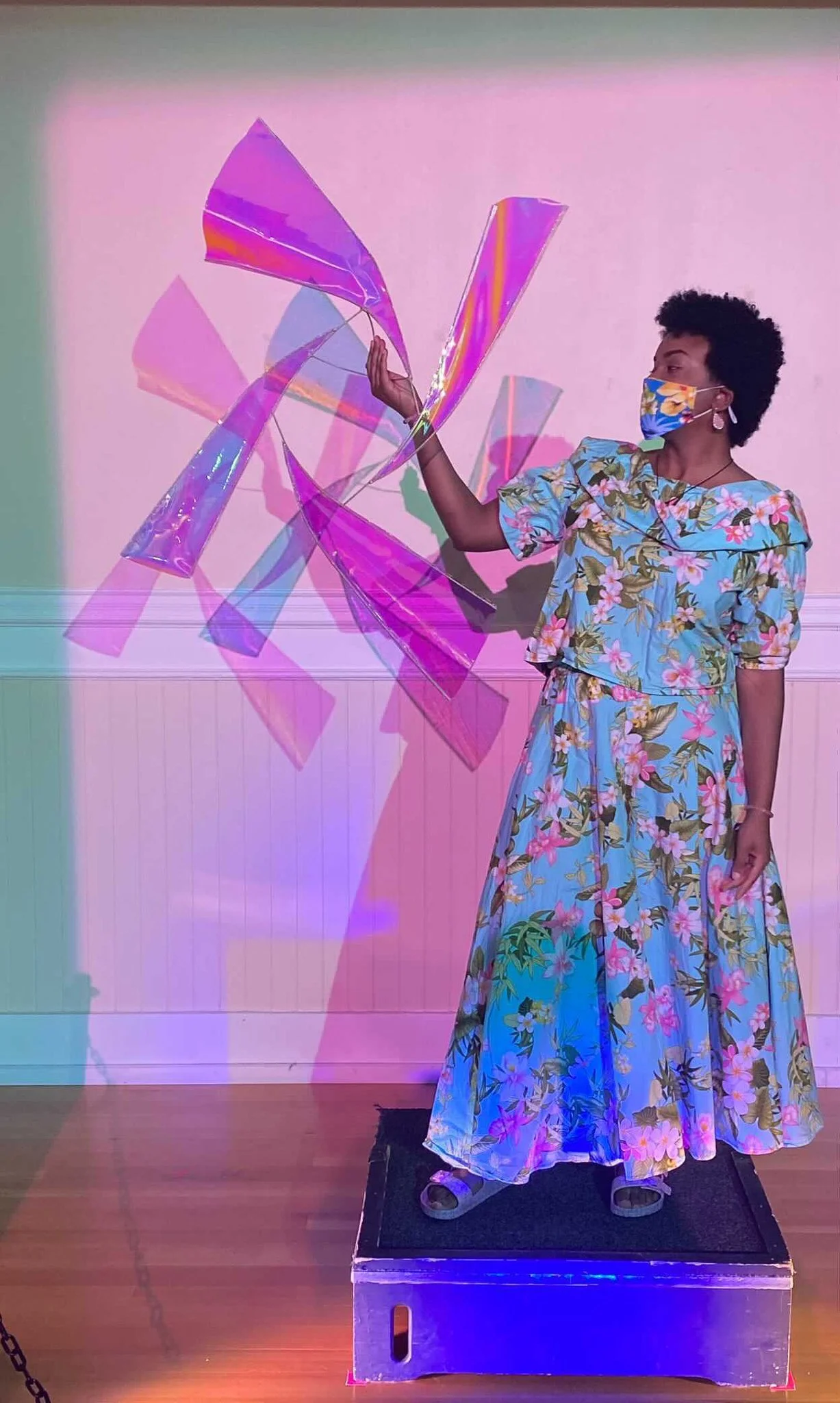 A black, female model wears a floral print with matching mask and holds a windmill sculpture extending from outstretched hand