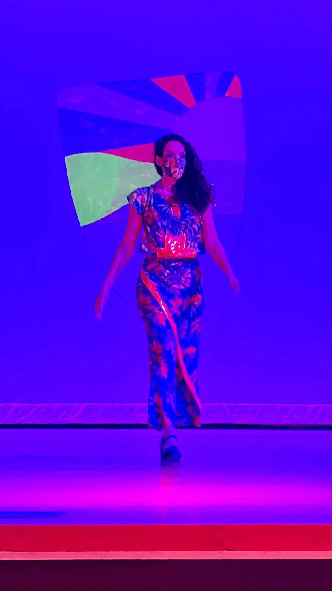 Black, female model wearing a luxuriously blue and orange patterned wrap dress on a neon glowing runway