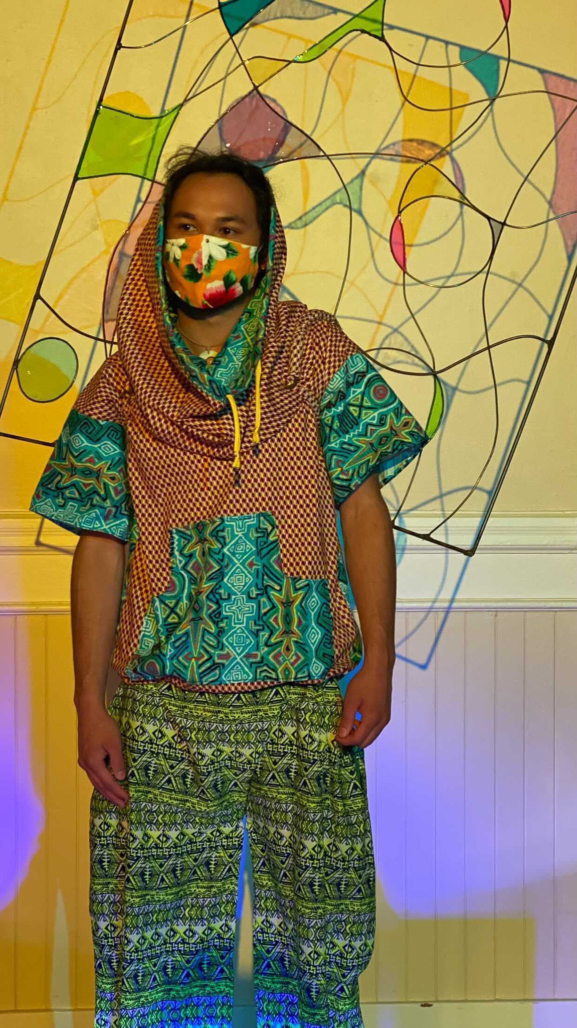 A black model wearing a patterned garment of teal and brown with a hoodie and colorful face mask. A delicate rectangular sculpture extends from his back.