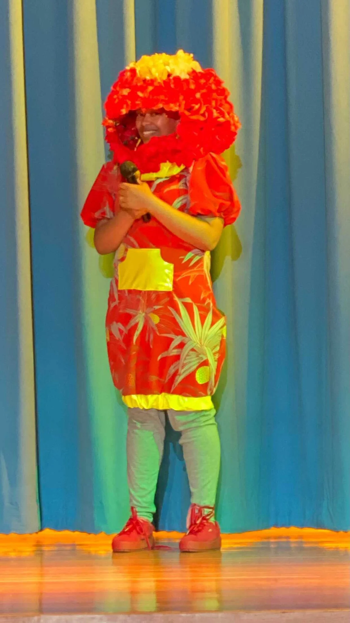 Afatasi the Artist wearing a red and yellow creation with an elaborate red and yellow flower head piece. She holds a microphone on stage.