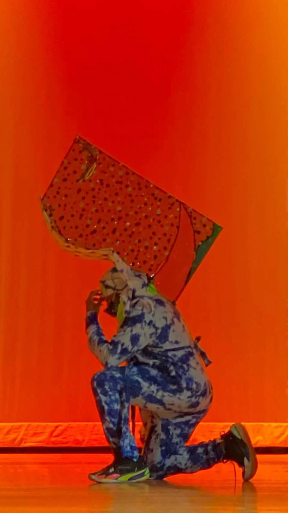 A male figure takes a knee on stage. We see him in profile. He wears a blue and white patterned track suit with matching hoodie and has a delicate shard sculpture extending from his back.