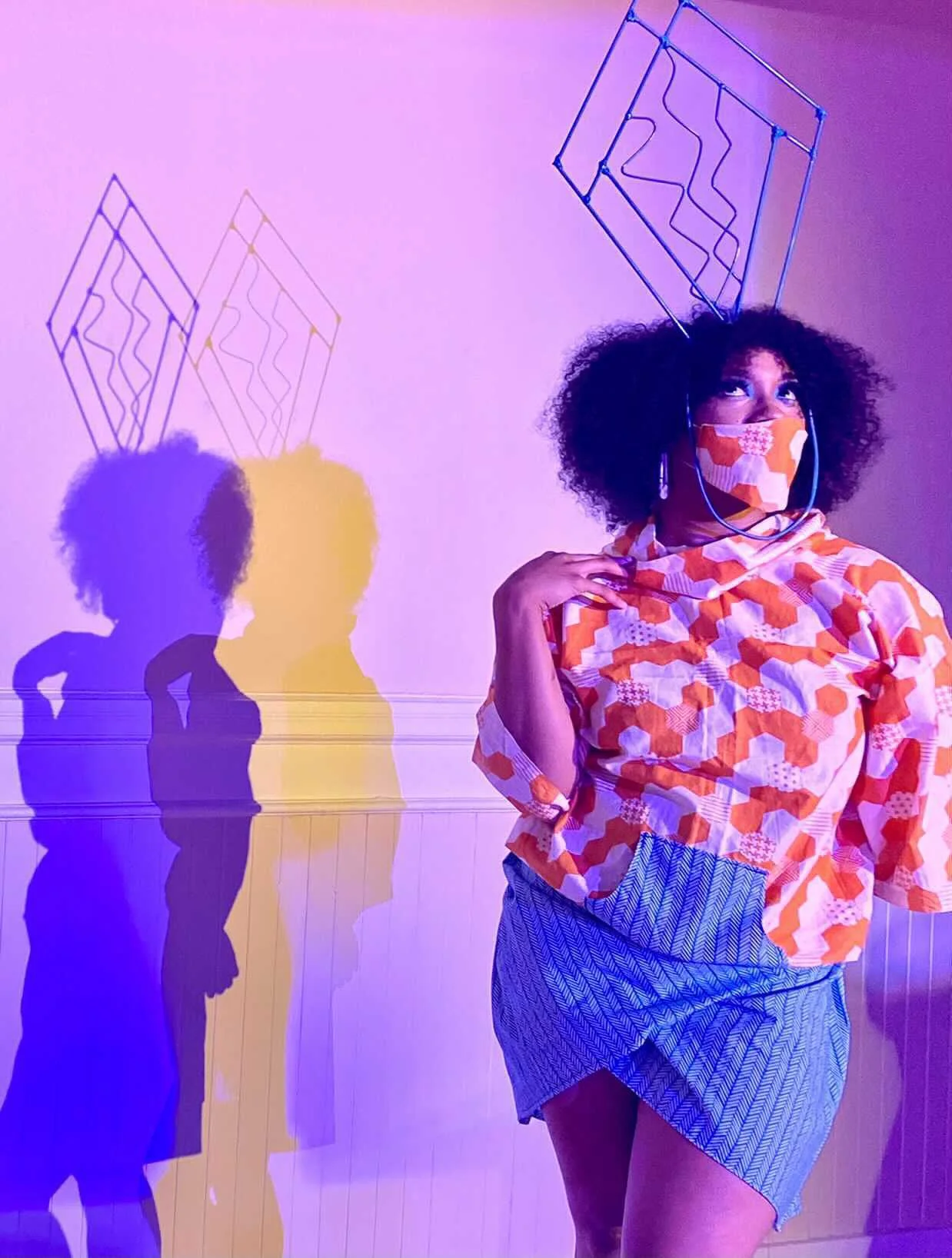 Black, female model wearing an orange patterned mask with matching top over a short blue wrap on a neon glowing runway