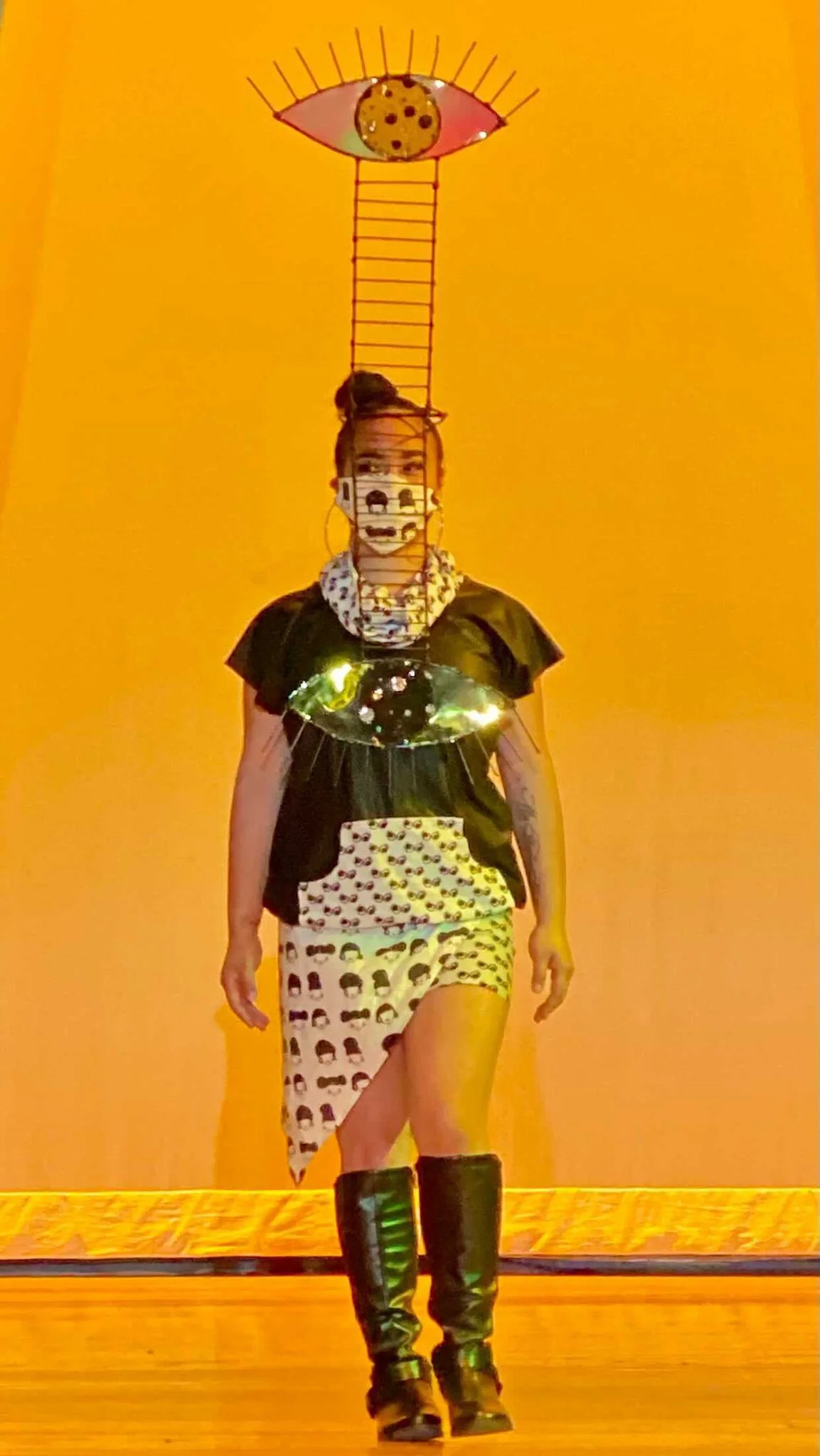 Black, female model wearing a black and white patterned, asymmetrical dress with a black breast plate and tall black boots. A ladder sculpture with an abstract eye on top extends from the top of her head