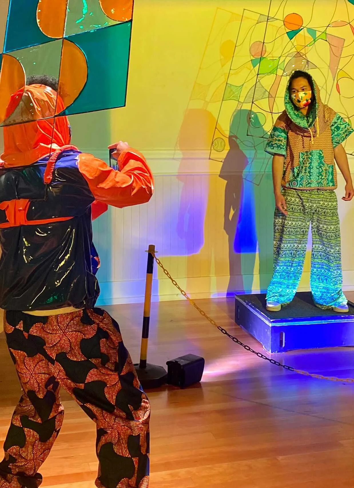 A model stands on a pedestal wearing a blue and tan patterned pants and matching top. Another person in orange and black snaps a photo. They both have colorful wire sculptures extending from their shoulders.