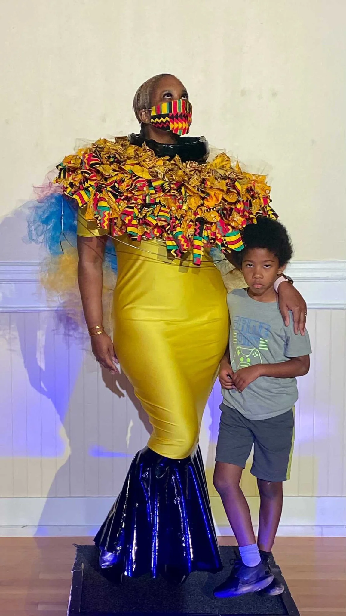 Black, female model wearing a  fitted, mermaid-style yellow dress with blue fins and an elaborate ruched collar. She wears a colorful mask and drapes her hand casually over the shoulder of a young, black boy standing beside her