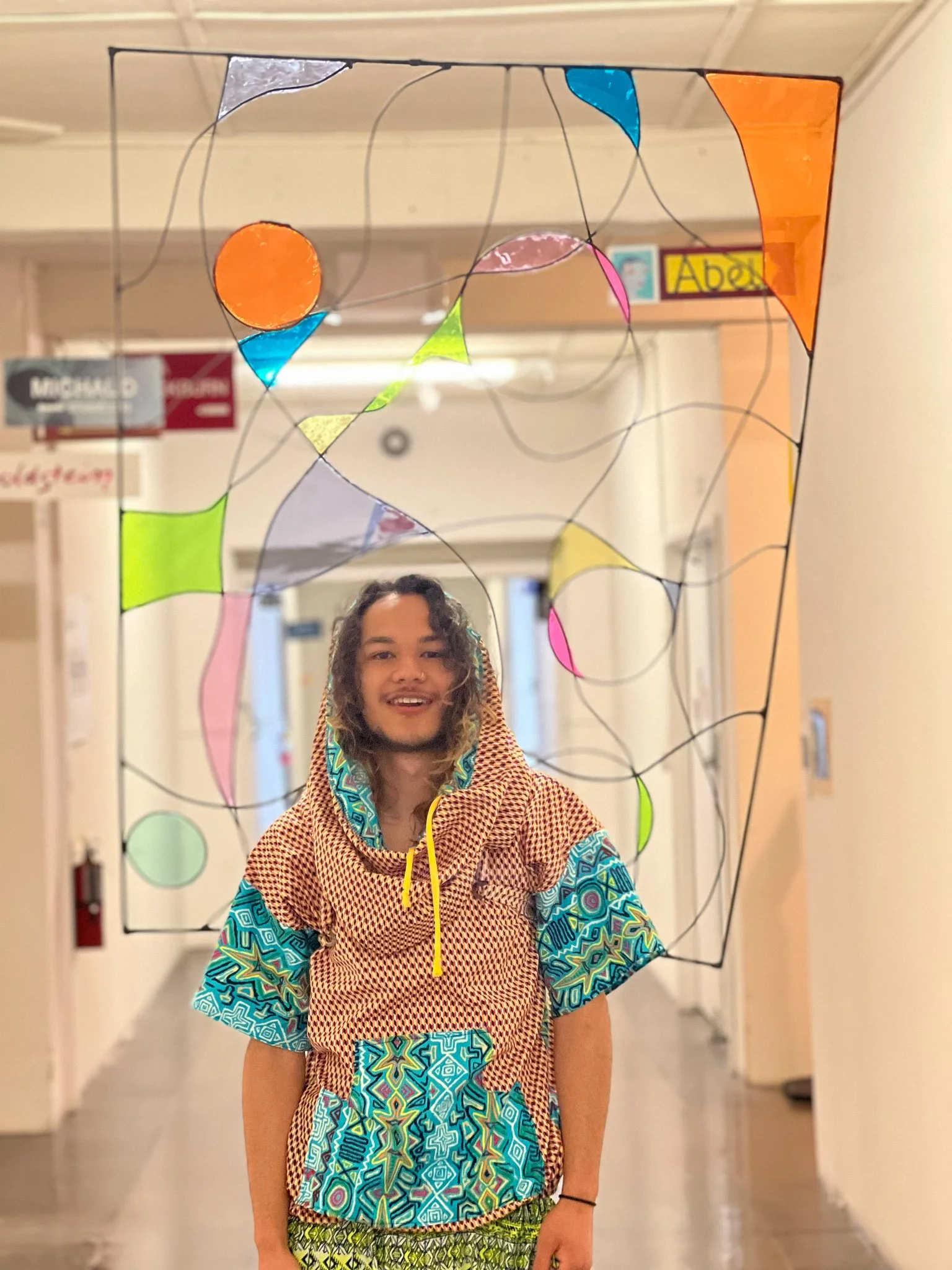 Black, male model wearing a blue patterned top with a hood standing in a long hallway with a large but delicate metal art piece behind him