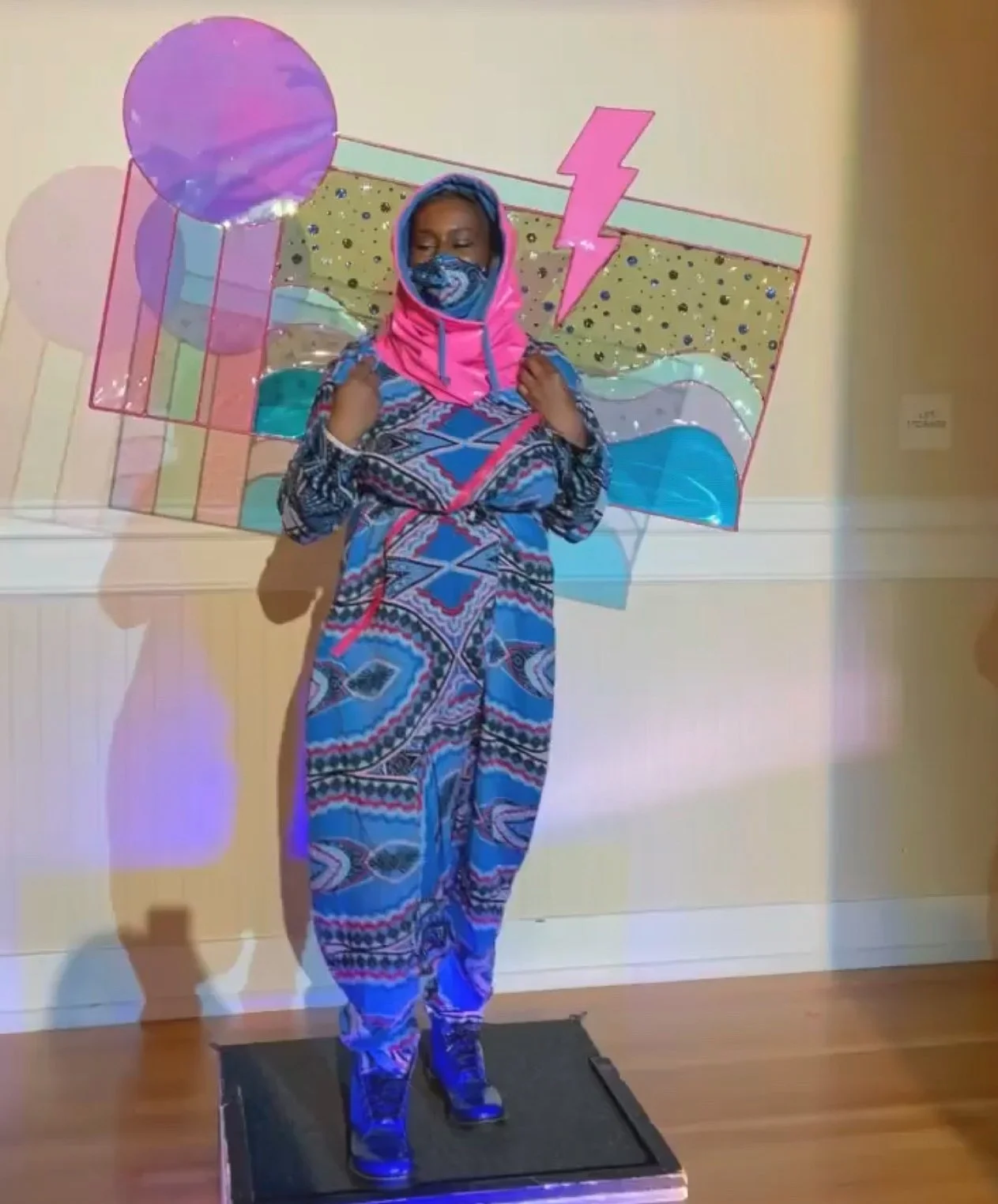 Black, female model wearing a blue and gray patterned jumpsuit with a matching face mask and a pink hoodie. She wears a delicate and colorful rectangular sculpture on her back.