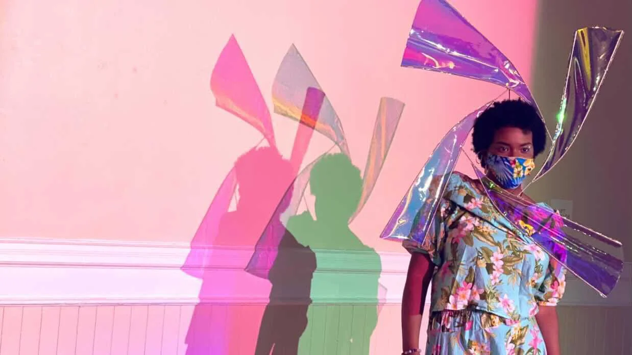 A black, female model wears a floral print with matching mask and has a delicate windmill sculpture extending from her back