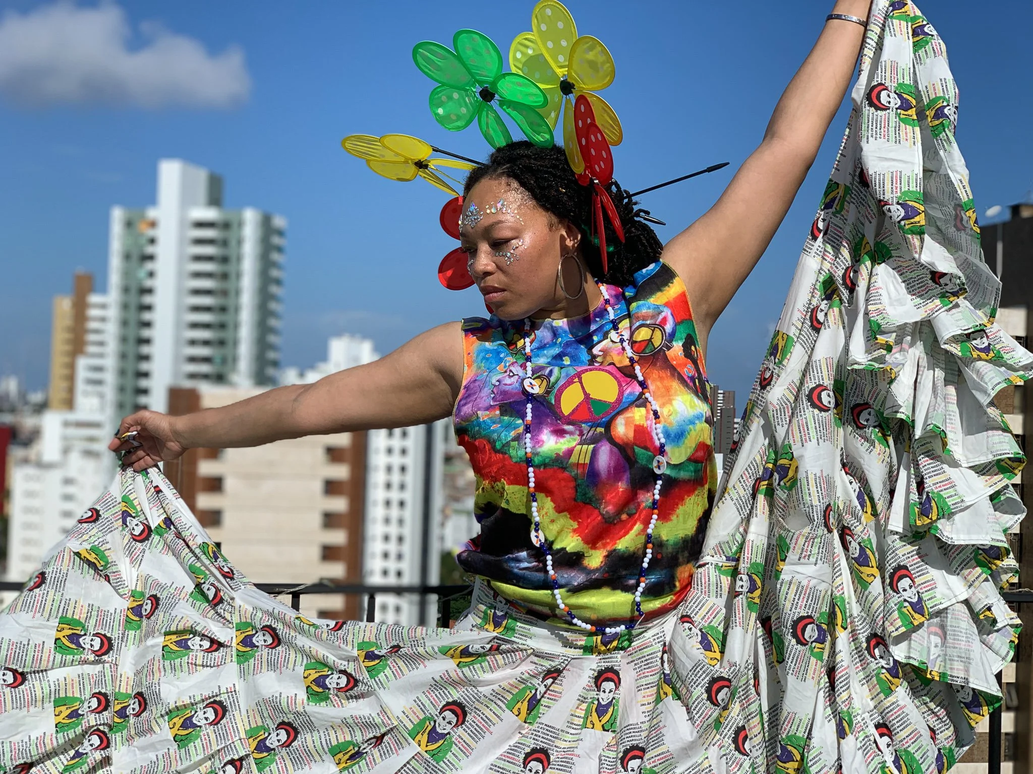 A black, female model stands in front of a cityscape. She wears a patterned skirt that she holds out to either side of her. She wears a rainbow tye dye tank top and has rainbow flowers in her hair