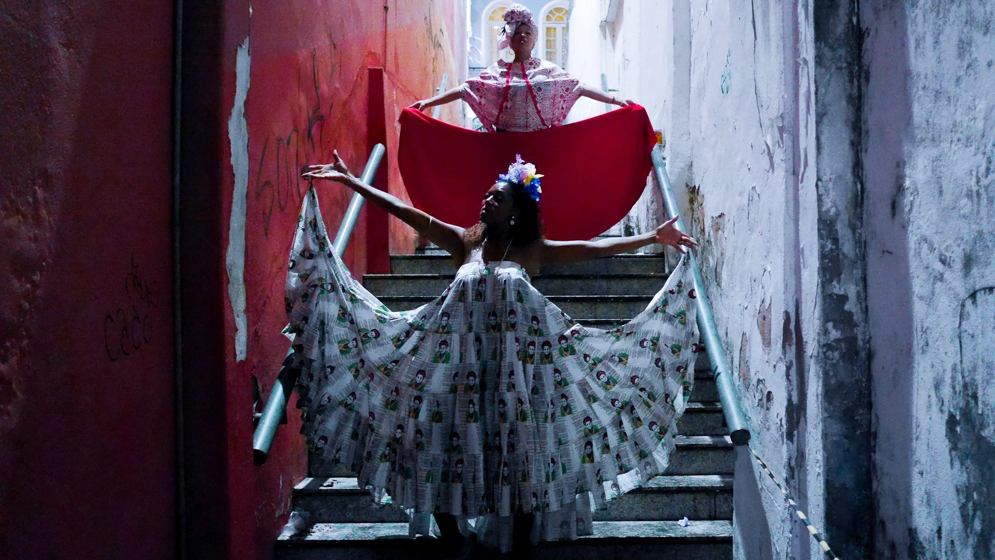 Two models on a staircase. The model in the foreground wears a silvery dress and the one in the background wears a red dress. They both hold their skirts out giving them an angelic presence.