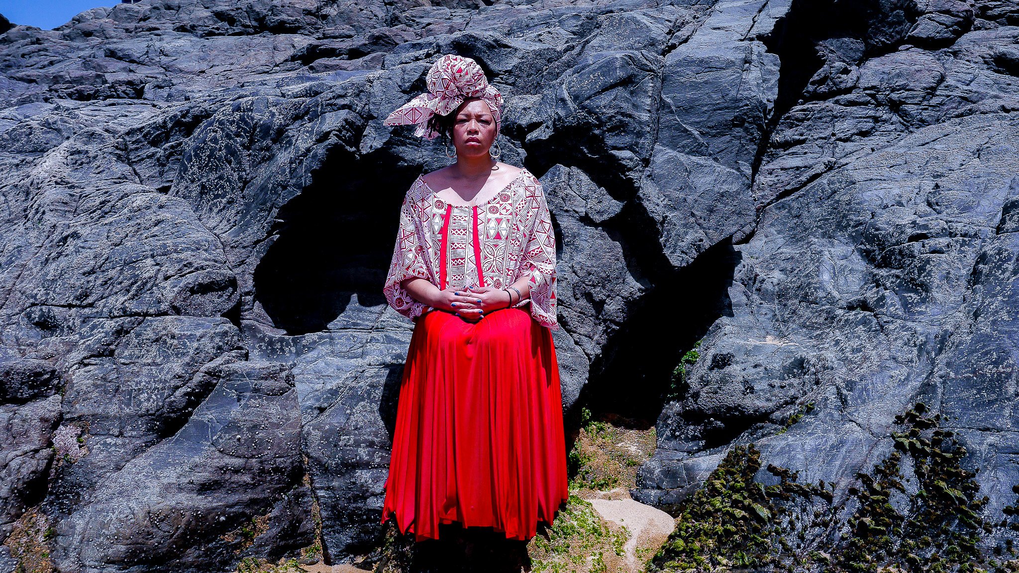 A black, female model stands in front of a rocky outcropping. She wears a long red skirt, and a white and red patterned top with matching head wrap.