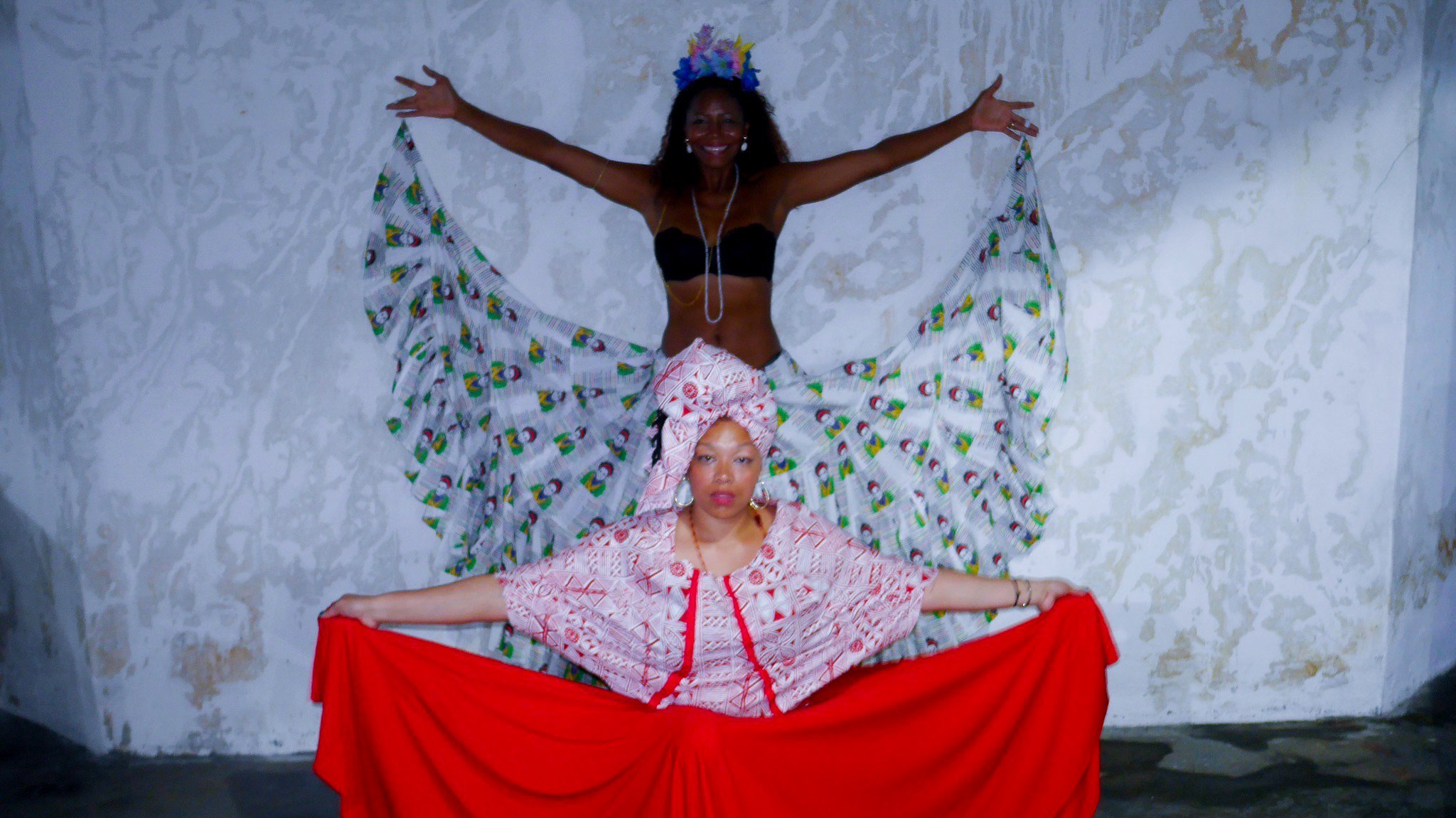 The model in the foreground wears a red dress and the one in the background wears a silvery skirt with a black bikini. top. They both hold their skirts out to their sides.
