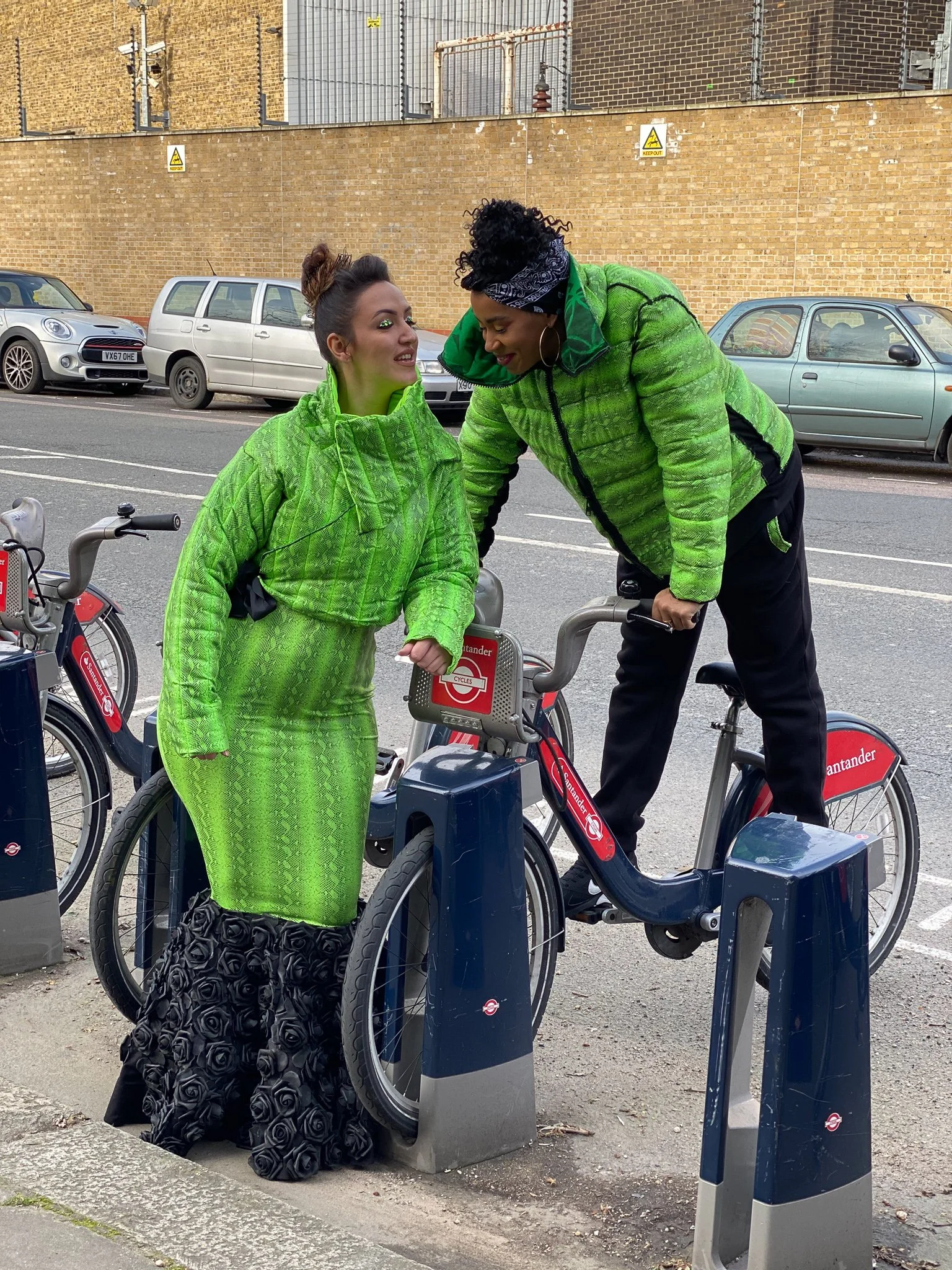 Two models. One wears a fitted green mermaid dress with black ruffling. The other wears a matching green jacket and black pants. They are posing at a rack of rental bicycles.