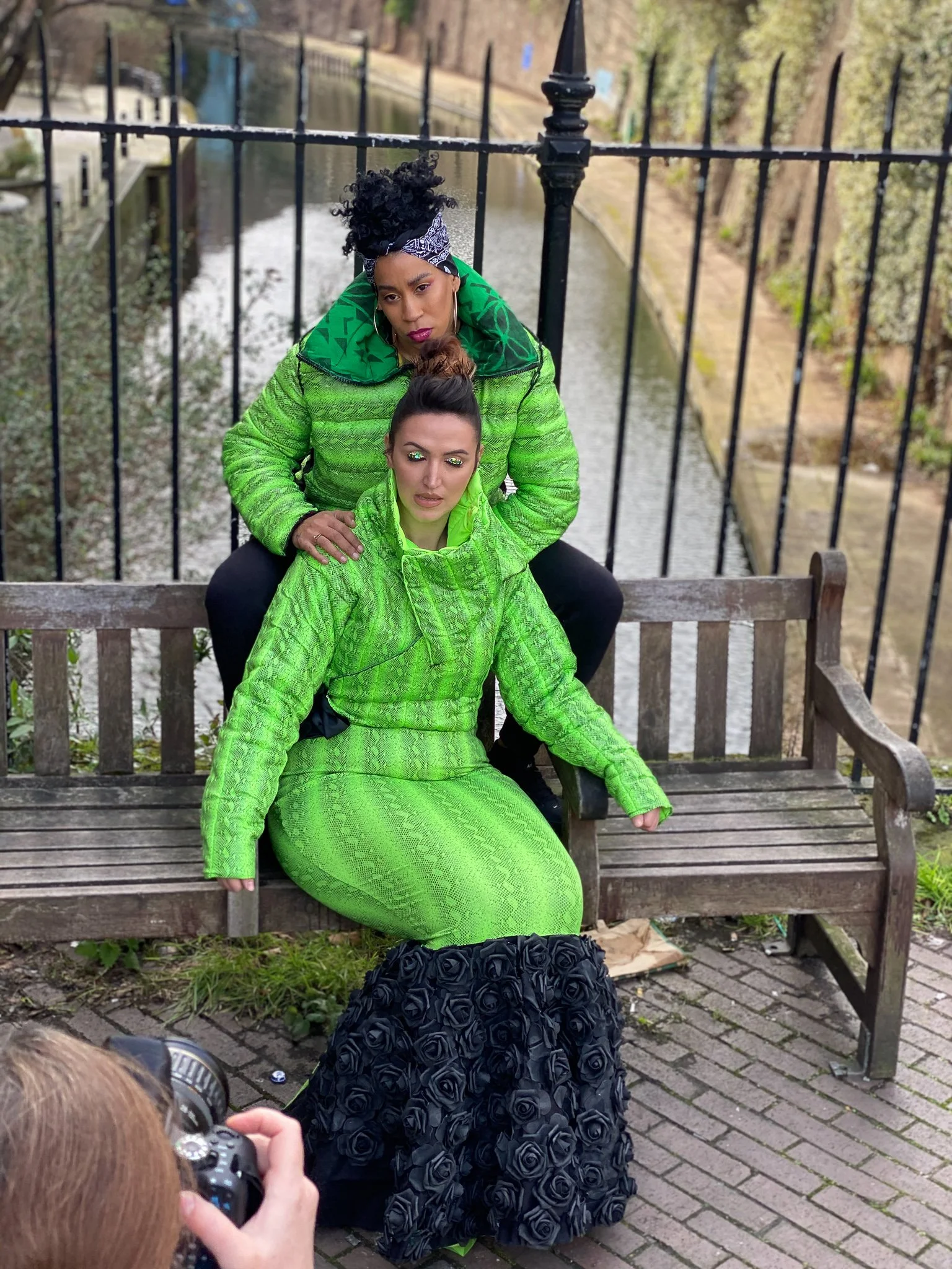 Two models. One wears a fitted green mermaid dress with black ruffling. The other wears a matching green jacket and black pants. They are seated together on a city bench.