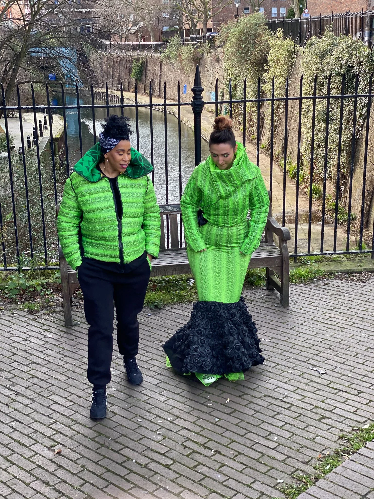 Two models. One wears a fitted green mermaid dress with black ruffling. The other wears a matching green jacket and black pants. They pose together in front of a city bench.