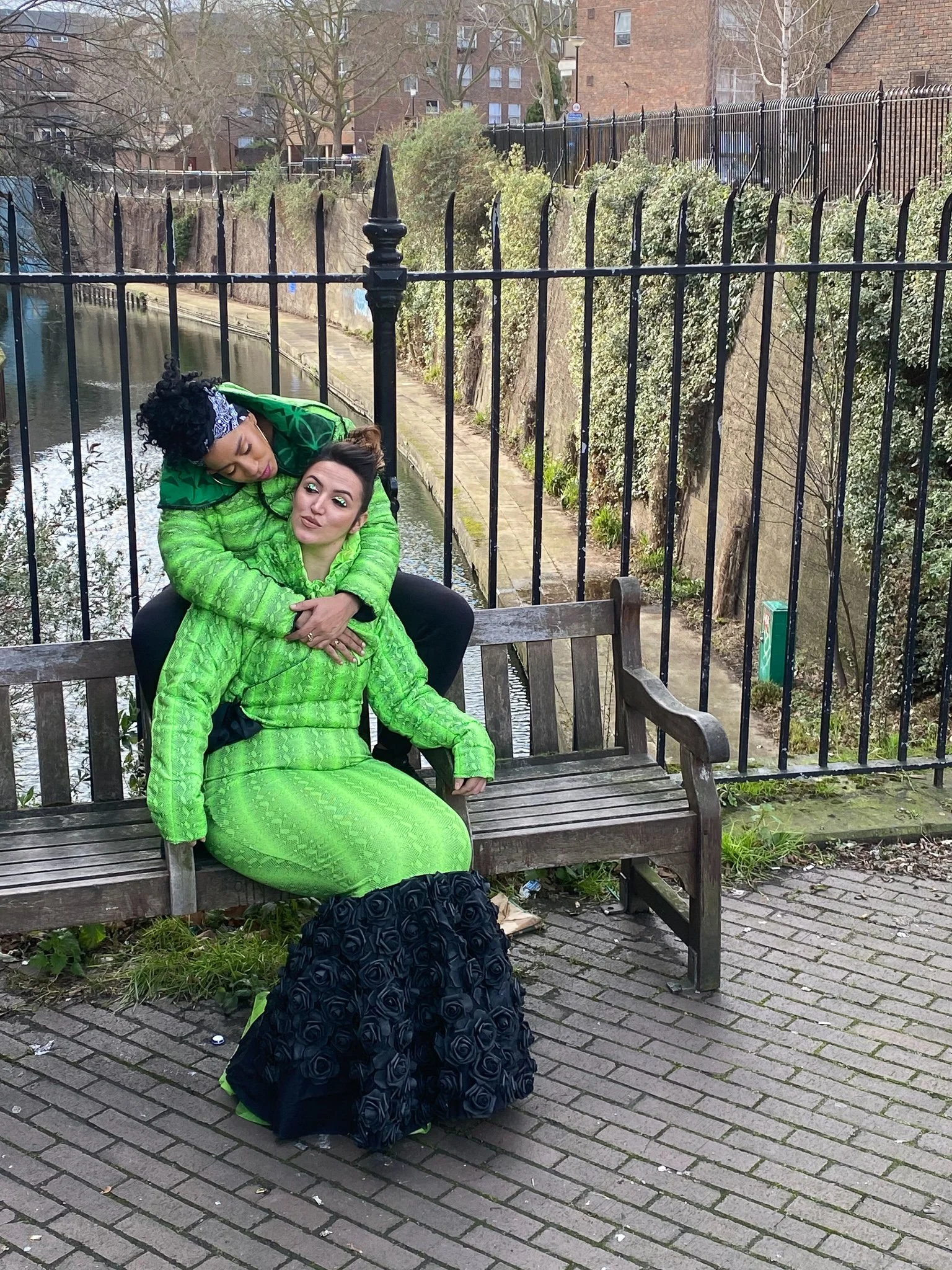 Two models. One wears a fitted green mermaid dress with black ruffling. The other wears a matching green jacket and black pants. They are seated together on a city bench with one draping their arms around the other.