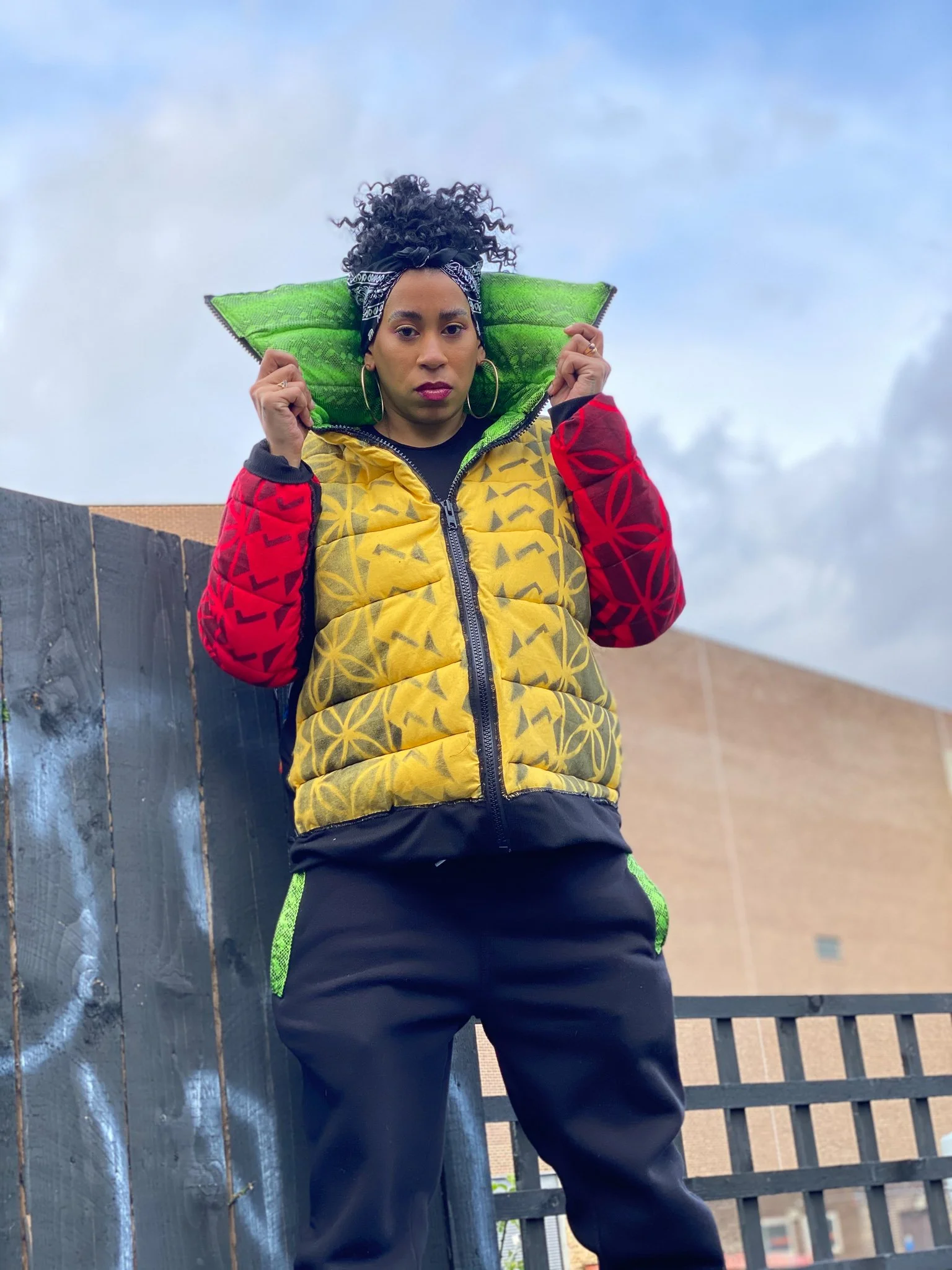 A black model posting in front of a graffiti wall. They wear a striking yellow, red, and green jacket with an exaggerated collar