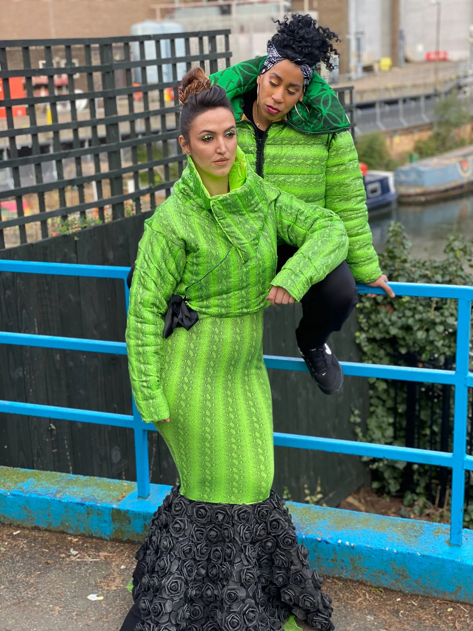 Two models. One wears a fitted green mermaid dress with black ruffling. The other wears a matching green jacket and black pants. They pose together against a blue city railing.