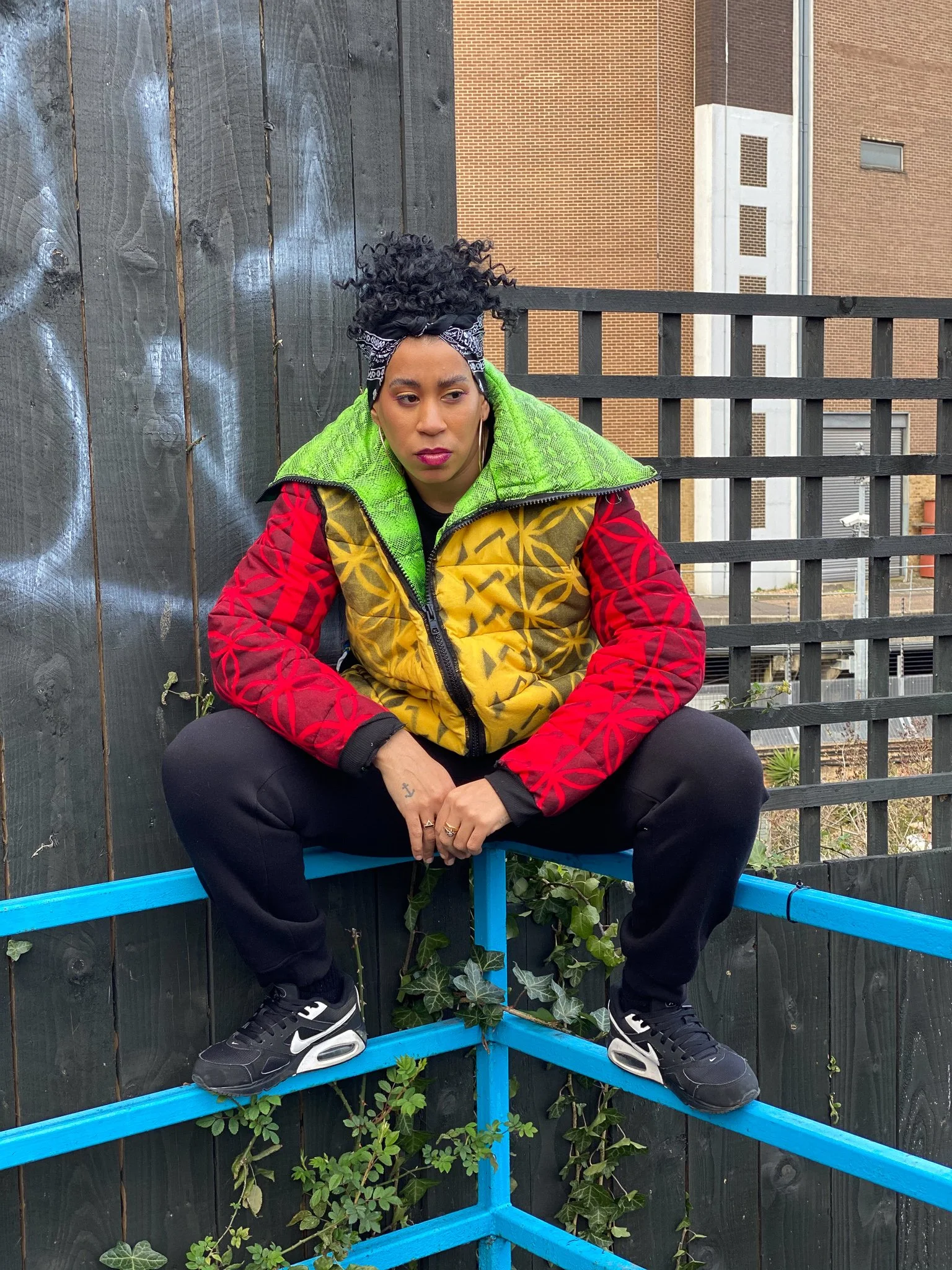A black model sitting on a blue, metal railing. They wear a striking yellow, red, and green jacket with an exaggerated collar