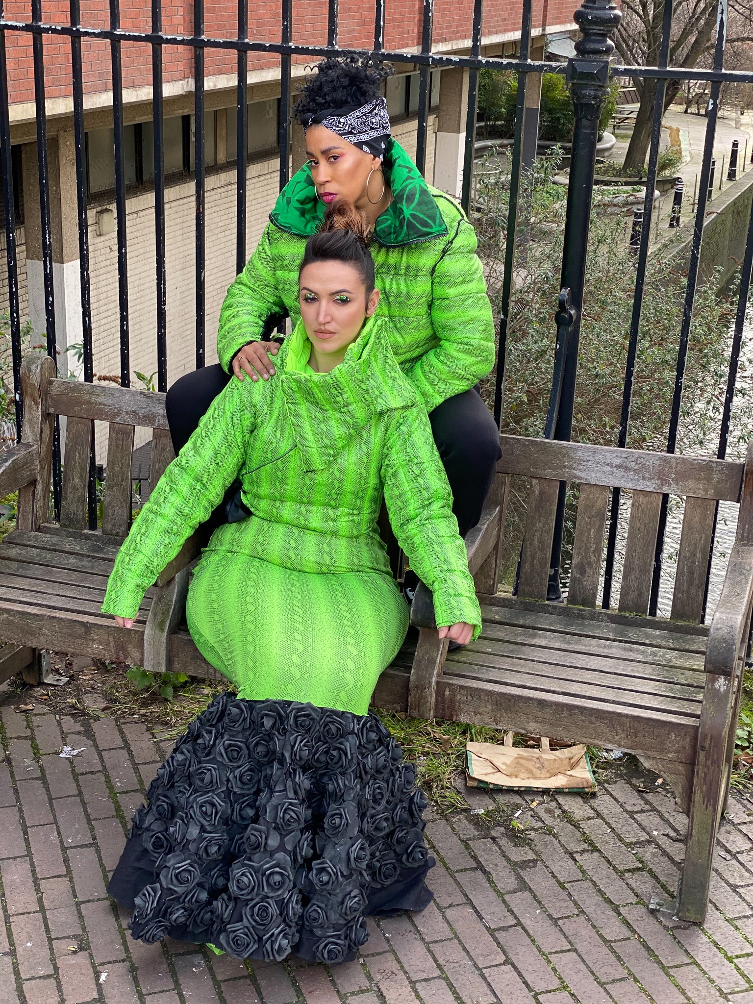 Two models. One wears a fitted green mermaid dress with black ruffling. The other wears a matching green jacket and black pants. They are seated together on a city bench.