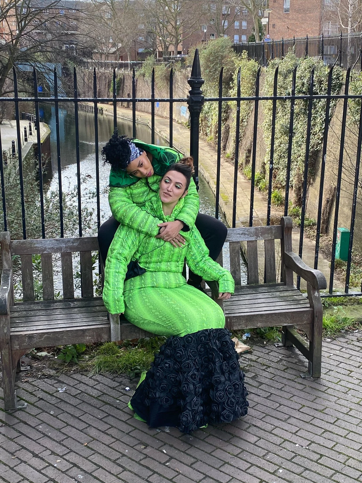 Two models. One wears a fitted green mermaid dress with black ruffling. The other wears a matching green jacket and black pants. They are seated together on a city bench with one draping their arms around the other.