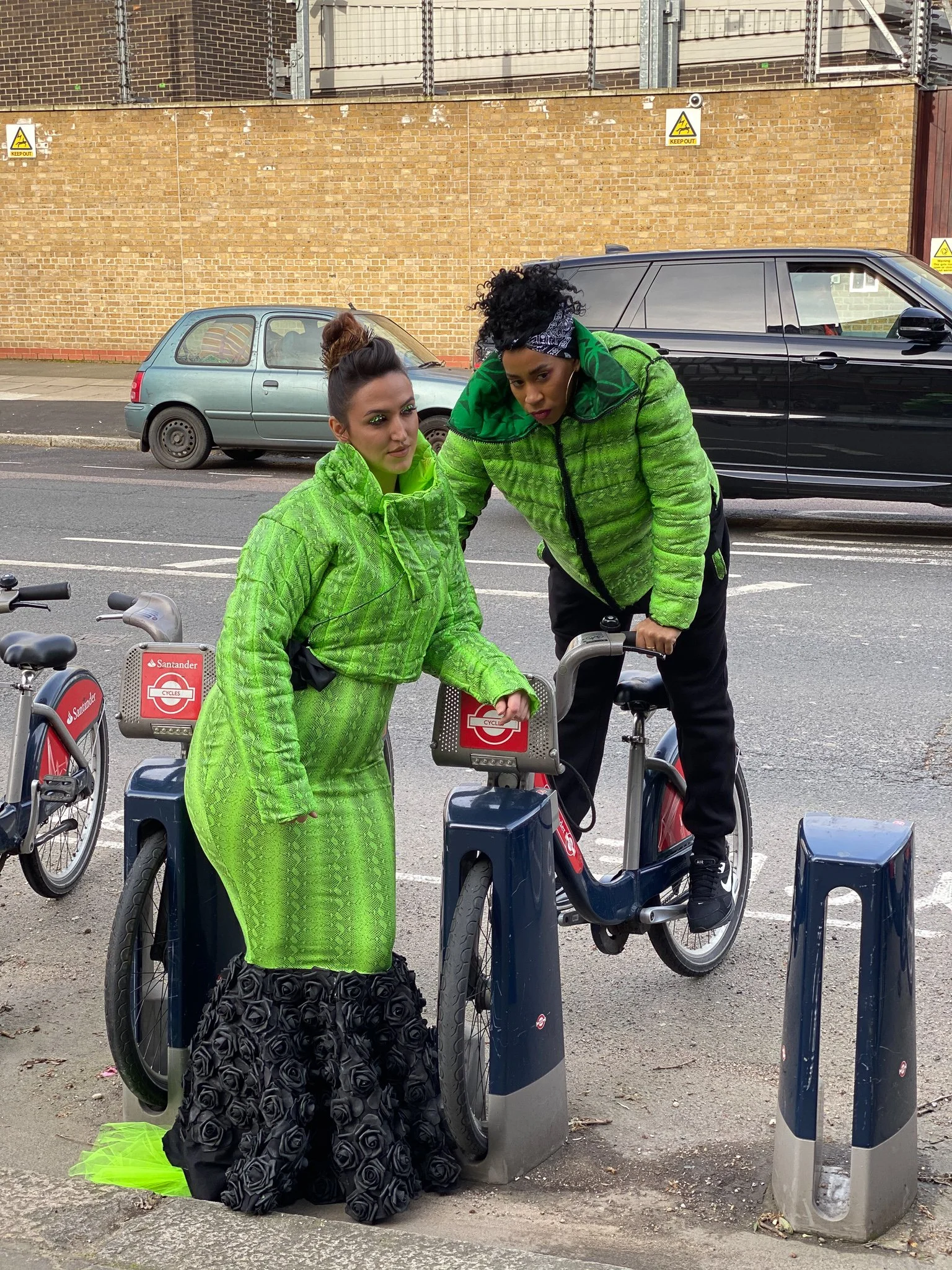 Two models. One wears a fitted green mermaid dress with black ruffling. The other wears a matching green jacket and black pants. They are posing at a rack of rental bicycles.