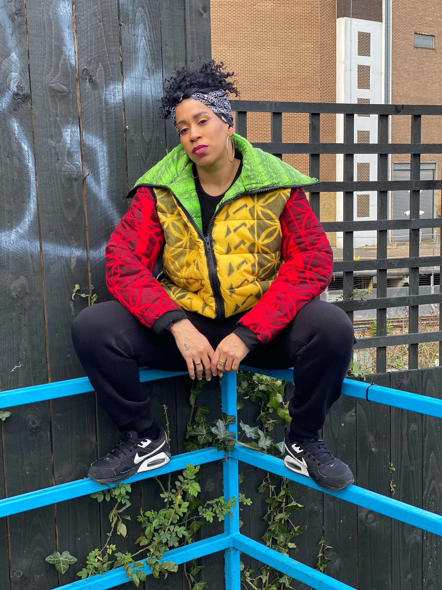 A black model sitting on a blue, metal railing. They wear a striking yellow, red, and green jacket with an exaggerated collar