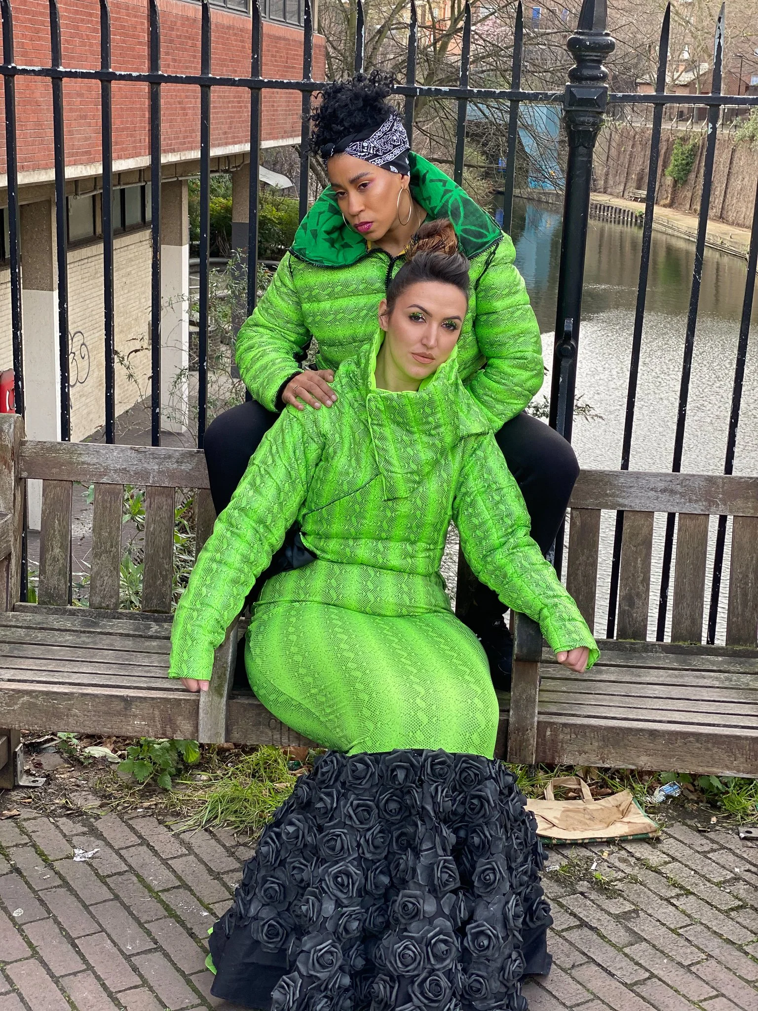 Two models. One wears a fitted green mermaid dress with black ruffling. The other wears a matching green jacket and black pants. They are seated together on a city bench with one resting their hands on the shoulder of the other.