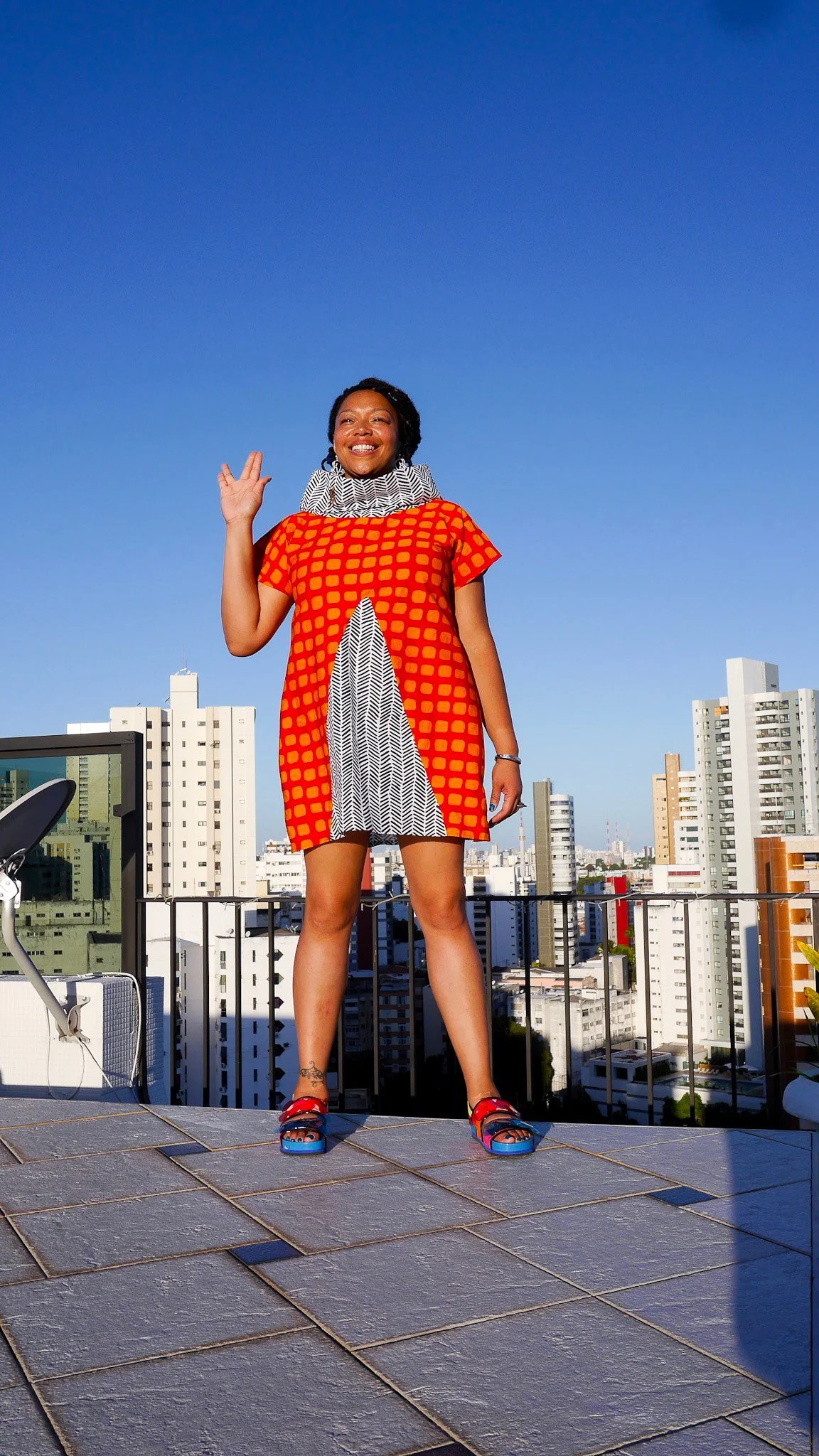 A model poses in front of a city scape  wearing a orange and gray garment with hand raised in a Spock greeting.