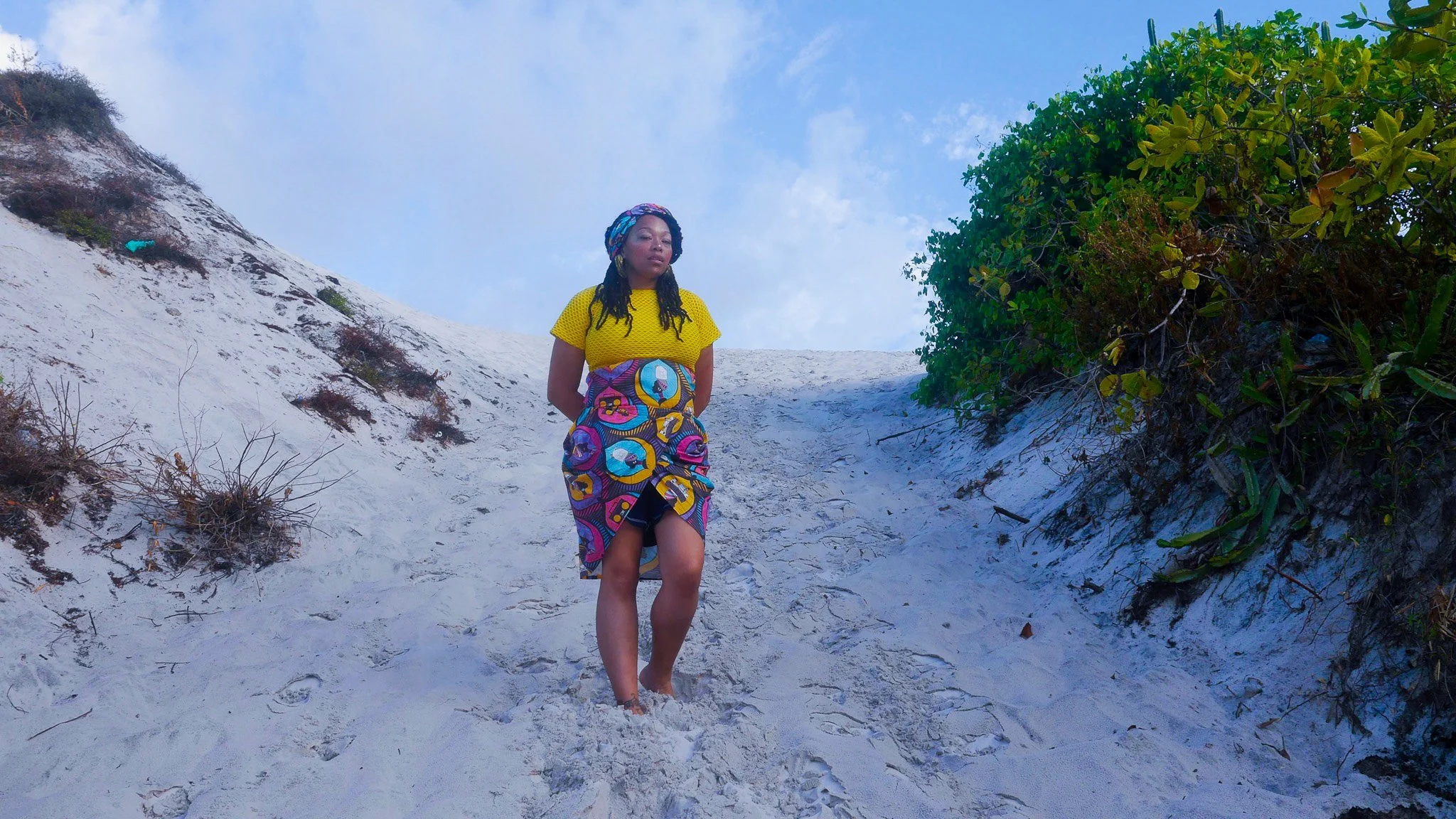 A black, female model stands on a sandy beach wearing a yellow top and a colorful patterned skirt that matches her head wrap