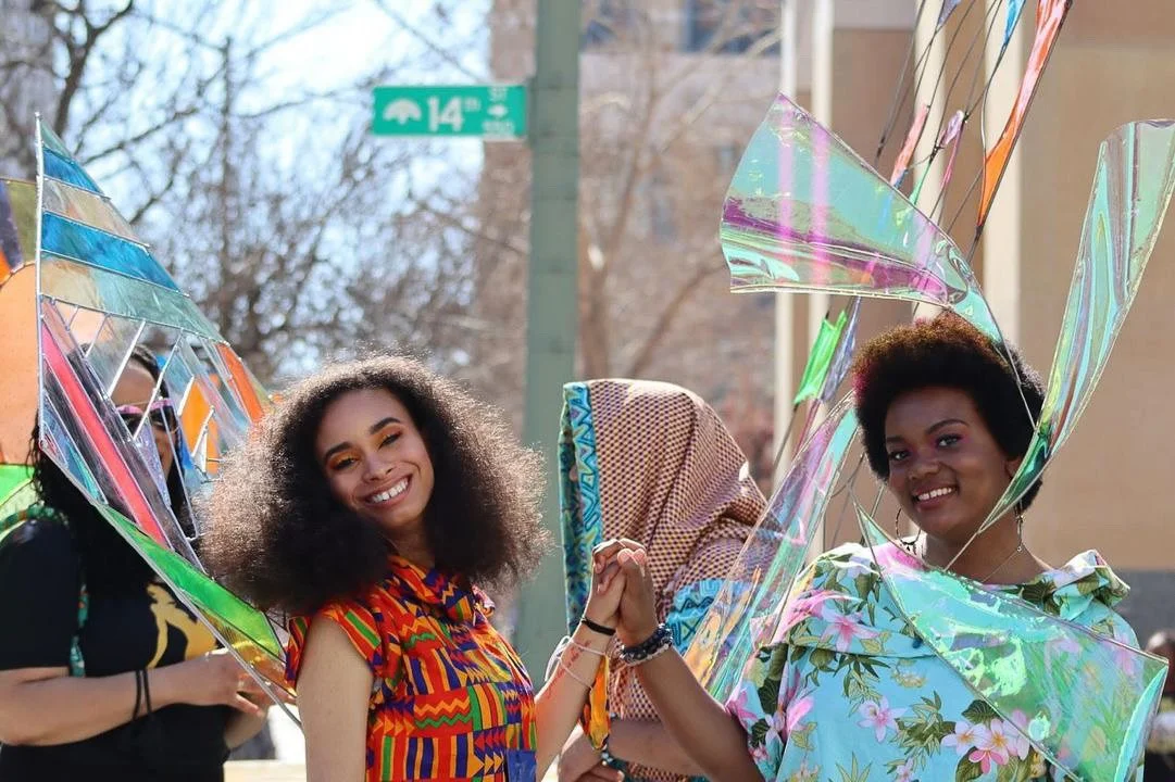 Two black, female models clasping hands outside in front of 14th street, Oakland. One wears an orange patterned top, the other  a blue floral print. They both have delicate metal sculptures extending from their backs.