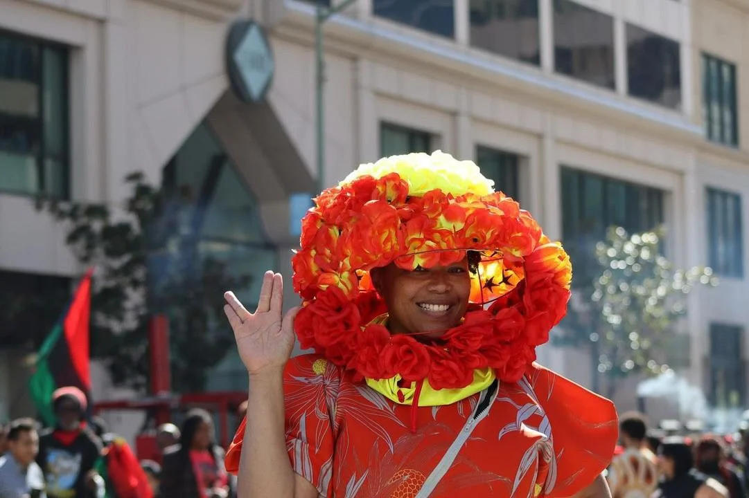 Afatasi the Artist stands outside a building, smiling with her hand raised in a Spock greeting.  She wears an elaborate red and yellow garment with a floral head piece.