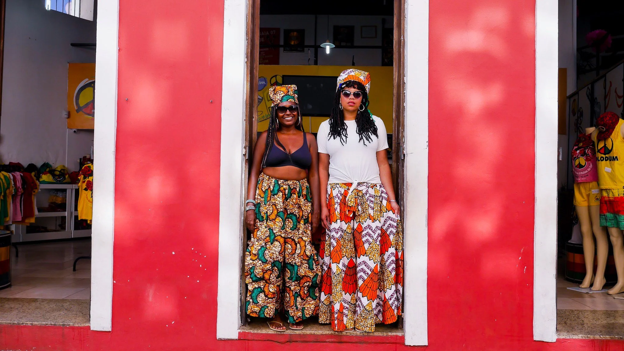 Two models stand in a red doorway. One wears a black bikini top and teal and gray and orange wide legged pants with matching head wrap. The other wears a white t-shirt, with tan, white, and orange wide legged pants and matching head wrap.