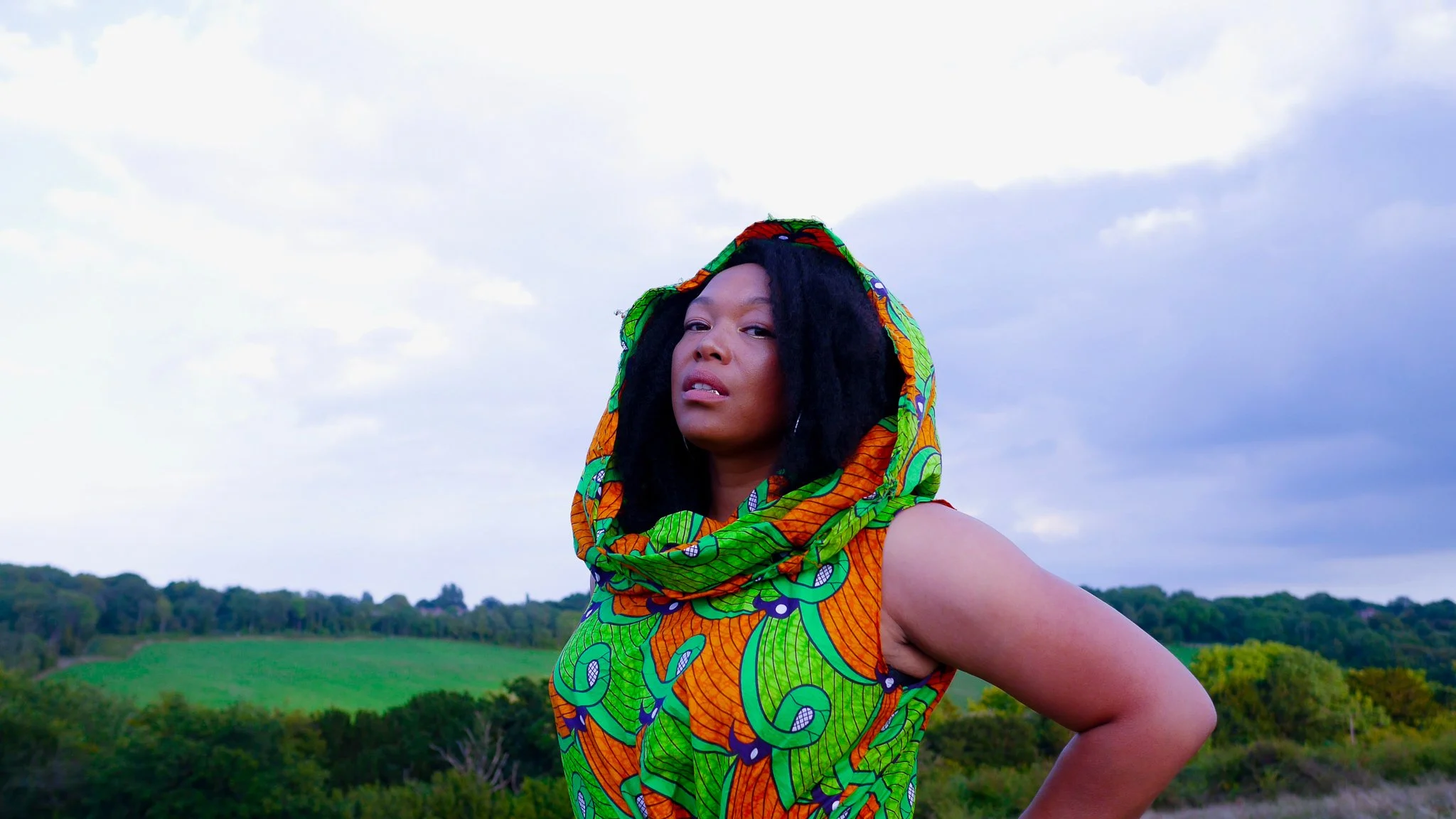 A black, female model poses in front of a lush green landscape wearing a green and orange print dress with matching hood.