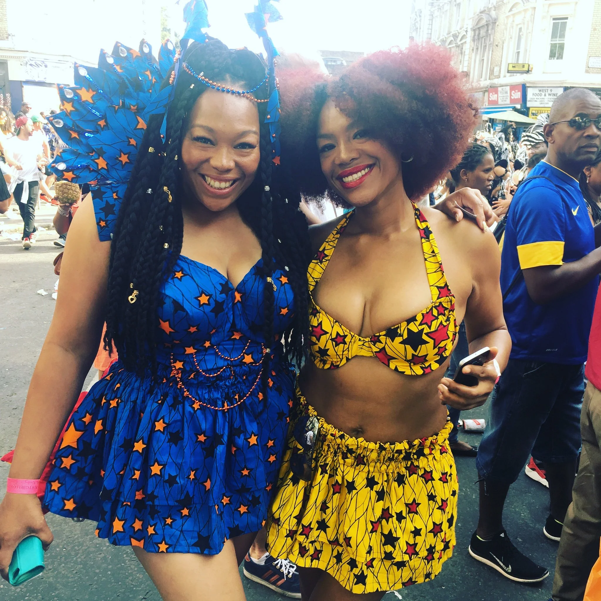 Two black, female models pose on a busy city street. One wears a blue and orange star dress with a matching head piece, the other wears a  similar yellow and red star patterned dress. 