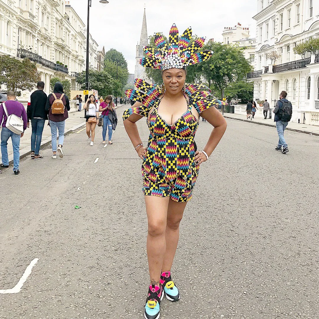 A model poses with a short brown, yellow, blue,  patterned dress with an elaborate matching head piece. She stands in the middle of a busy city street with bright neon sneakers.
