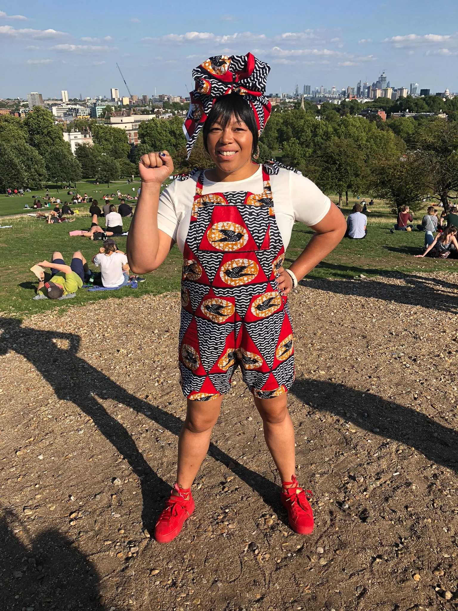 A black model poses in front of a city park wearing a gray, orange, and red overall shorts with matching head wrap and red sneakers.