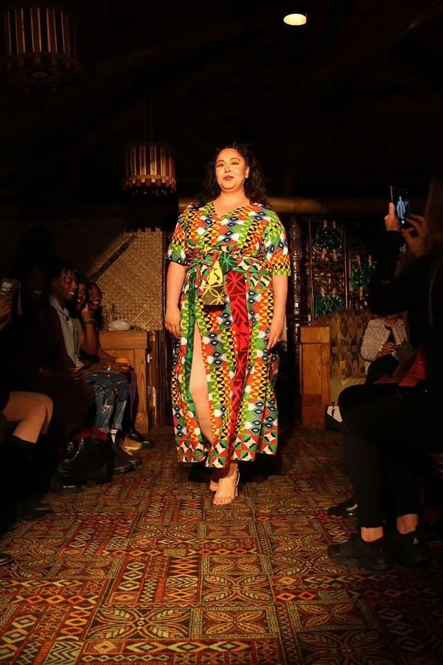 A model walks the runway wearing a flowing green, yellow, orange, and red dress.