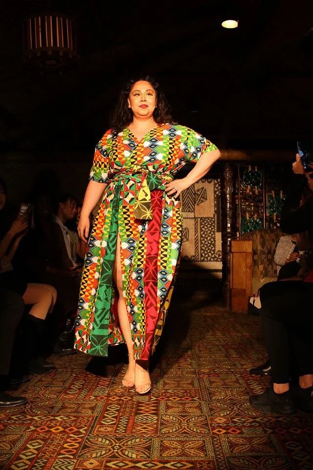 A model walks the runway wearing a flowing green, yellow, orange, and red dress.