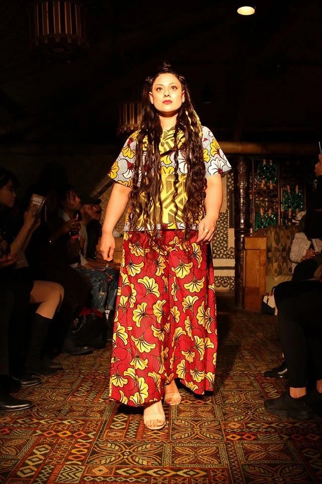 A model walks the runway wearing a flowing red and yellow patterned skirt with a yellow and white patterned top.