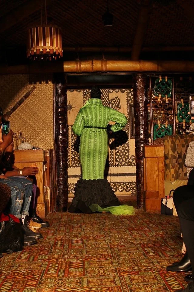 A black, female model exits the runway wearing a fitted, green mermaid dress with black ruffles.