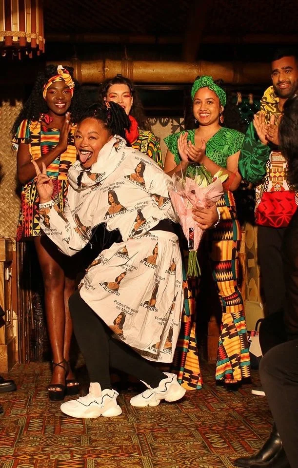 A group of three models stand together in colorful garments.  Afatasi the Artist poses jubilantly in the foreground with flowers in her hand.